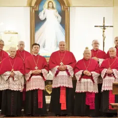 Grupa duchownych w strojach liturgicznych, w tym kilku kanoników, pozuje do zdjęcia przed obrazem Jezusa Miłosiernego.