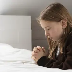 Dziewczynka z zamkniętymi oczami, składając dłonie, wykonuje akt żalu dla dzieci. Modli się na białej pościeli.