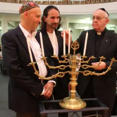 Trzech mężczyzn, jeden w jarmułce, zapala świece na menorze.