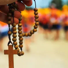 Drewniany różaniec z krzyżem w dłoni, symbolizujący modlitwę za brata. W tle rozmyte sylwetki ludzi.