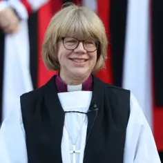 Uśmiechnięta kobieta w stroju duchownym, z krzyżem na szyi, przypomina arcybiskupa Canterbury.