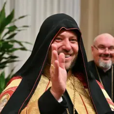 Uśmiechnięty, najmłodszy kardynał w bogato zdobionej szacie wita. W tle inny duchowny.