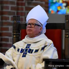 Biskup w białej infule i ozdobnej szacie siedzi na drewnianym krześle.