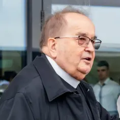 Ksiądz Rydzyk w okularach i czarnej kurtce, z białym kołnierzykiem, wychodzi na zewnątrz.