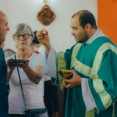 Kapłan udziela komunii, przypominając o prawdzie wiary. Kobieta z naczyniem przyjmuje hostię.