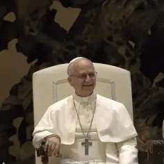 Uśmiechnięty Ojciec Święty w białej szacie, z krzyżem na szyi, siedzi na tronie. Obok niego stoi duchowny. To chwila modlitwy do Ojca Świętego.