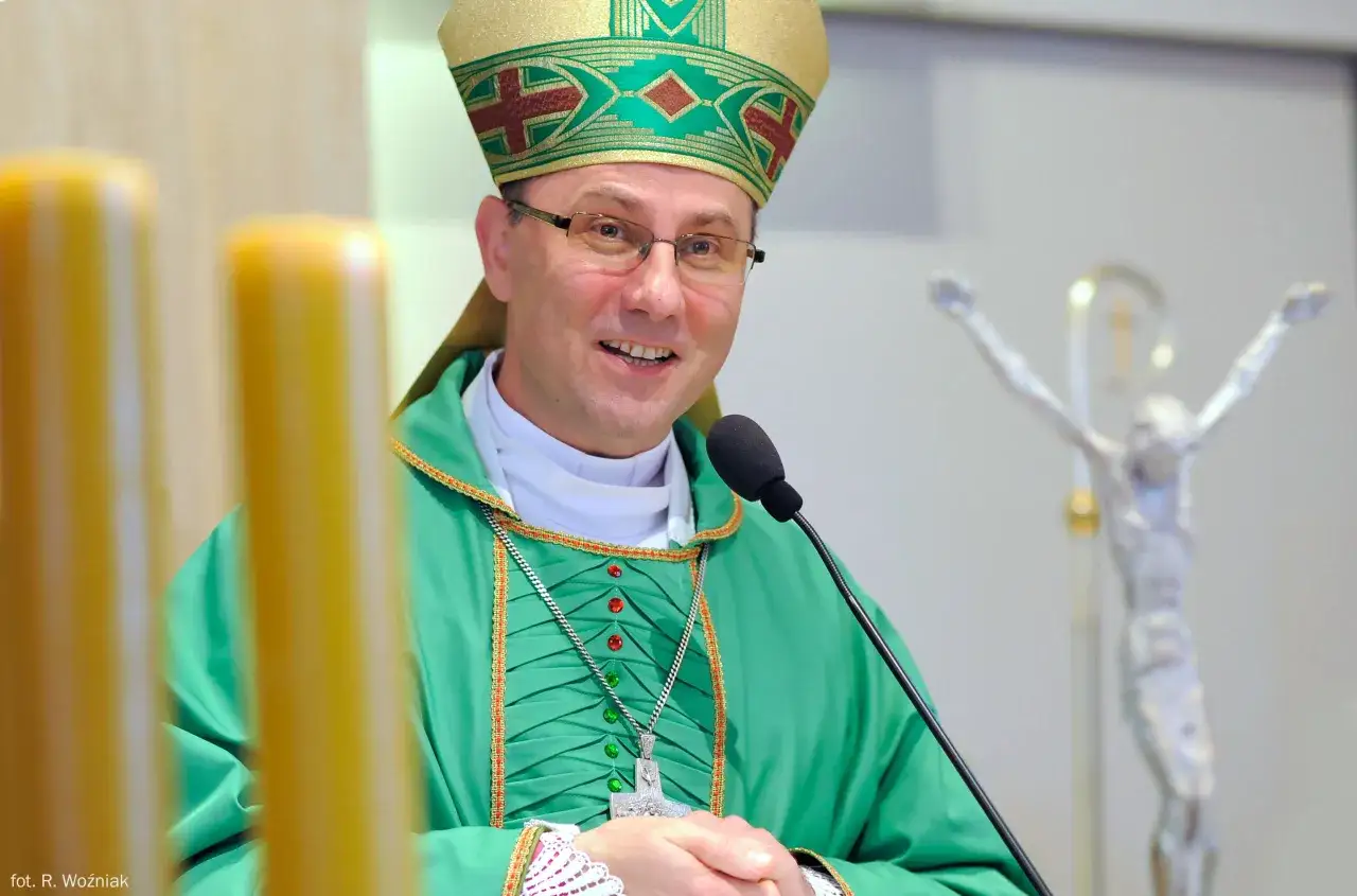 Uśmiechnięty prymas polski w zielonej szacie i mitrze, przed mikrofonem, obok świec i krucyfiksu.