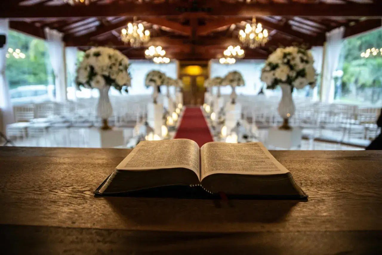 Otwarta Biblia na drewnianym stole, symbol aktu wiary. W tle ozdobiona sala z krzesłami i kwiatami.